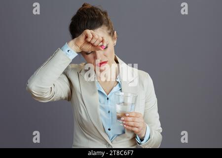 Müde elegante 40 Jahre alte Arbeiterin in einem leichten Business-Anzug mit Glas Wasser und Pille isoliert auf grauem Hintergrund. Stockfoto