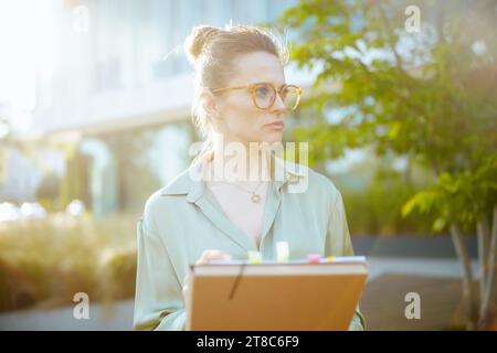 Nachdenkliche, moderne Geschäftsfrau mittleren Alters in der Nähe des Bürogebäudes in grüner Bluse und Brille mit Dokumenten und Ordner. Stockfoto