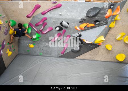 Junger Mann klettert auf die Trainingswand im Sportzentrum. Starker erfahrener Rock Climber, der im Fitnessstudio Solo-Klettern an der Boulderwand übt. Mann, der im Fitnessstudio trainieren kann. Aufnahme von hinten Stockfoto