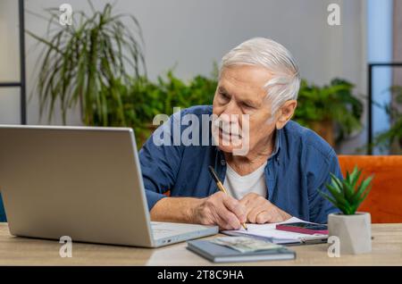 Senior man Buchhaltungszahlungen berechnen monatliche Ausgaben Bankrechnungen analysieren Papierkram in der Wohnung drinnen. Älterer, reifer Großvater, freiberuflicher Online-Job auf Laptop am Tisch Stockfoto