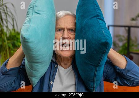 Reparaturarbeiten bei Nachbarn. Irritierte ältere Großvater Mann schreiend Decke Ohren mit Kissen, genervt von lauter Partymusik wünscht Stille. Dünne Wände im Wohnbereich ohne Schalldämmung Stockfoto