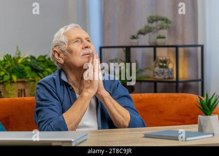 Bitte, Gott helfe. Besorgter Senior Mann, der aufrichtig um Heide betet, mit gefalteten Armen, die nach oben schauen, was sich wünscht, und bittet um hoffnungsvollen, bittenden Ausdruck, der sich zu Hause um Entschuldigung bittet. Der alte kaukasische Großvater Stockfoto