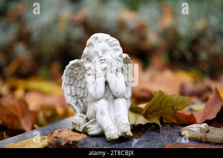 Eine kleine weiße Engelsfigur sitzt träumend auf einem Grabstein mit Herbstlaub Stockfoto