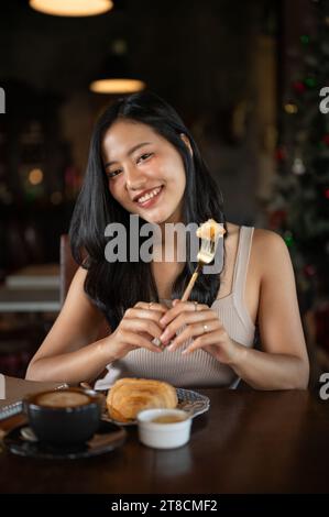 Elegante junge asiatische Frau, die Croissant zum Frühstück mit heißem Latte in einem dunklen luxuriösen Wohnzimmer isst. Morgens Frühstück mit Kaffee. Stockfoto