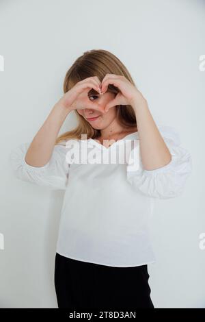 Eine Frau in weiß machte ein Herz aus ihren Fingern und schaut hinein Stockfoto