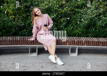 Blonde Frau, die auf einer Bank sitzt und eine Tasse Drink an den grünen Büschen trinkt Stockfoto