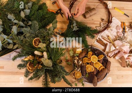 Draufsicht Komposition weiblicher Hände, die Weihnachtskranz aus natürlichen Zweigen von Fichte, Kiefer, Eukalyptus herstellen. Das Mädchen wickelt grüne Zweige auf die Kraft-Basis Stockfoto