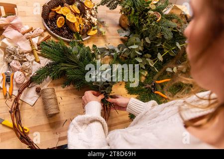 Draufsicht Komposition weiblicher Hände, die Weihnachtskranz aus natürlichen Zweigen von Fichte, Kiefer, Eukalyptus herstellen. Das Mädchen wickelt grüne Zweige auf die Kraft-Basis Stockfoto