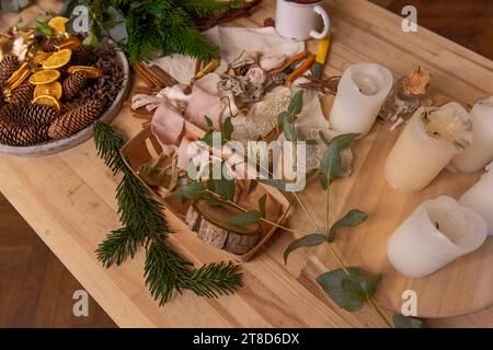 Auf dem Holztisch befinden sich rustikale Materialien für die kreative Dekoration der Weihnachtseinrichtung. Tannenzweige weiße Kerzen, Eukalyptuskieferkegel getrocknete Orangen. W Stockfoto