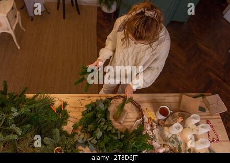 Blick von oben auf die junge Frau, die zu Hause aus natürlichen Materialien handgefertigte Weihnachtskränze herstellt. Wohndekoration für Silvesterferien. Blogger schreibt DIY-ins Stockfoto