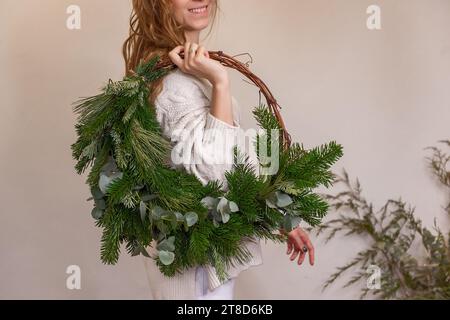 Gesichtslose junge Frau hält asymmetrischen grünen Weihnachtskranz aus natürlichen Materialien in den Händen. Mädchen in gestricktem beigefarbenem Pullover macht Dekorationen mit Stockfoto