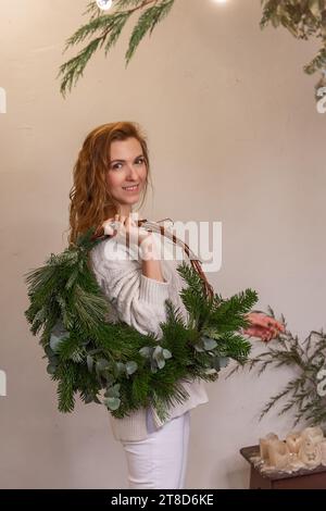 Porträt einer jungen Frau mit asymmetrischem grünen Weihnachtskranz aus Naturmaterialien auf der Schulter in den Händen. Mädchen in gestricktem Beige mit Homemad Stockfoto