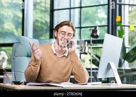 Glücklicher Mann im Büro, der am Telefon spricht, am Tisch vor dem Computer sitzt, ein erfolgreiches Geschäftsgespräch führt und mit den Händen Gesten macht. Stockfoto