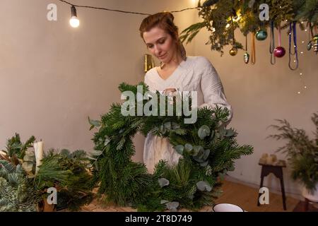 Offene junge Frau im Strickpullover macht flauschigen, grünen Weihnachtskranz aus natürlichen Fichten-, Kiefer- und Eukalyptuszweigen. Warme, winterliche Atmosphäre Stockfoto