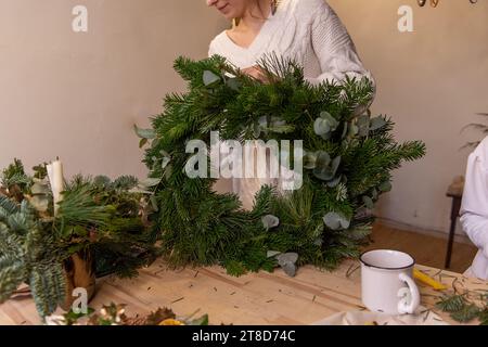 Faceless young woman in knitted sweater makes fluffy, green Christmas wreath from natural spruce pine, eucalyptus branches. Warm, winter atmosphere in Stockfoto