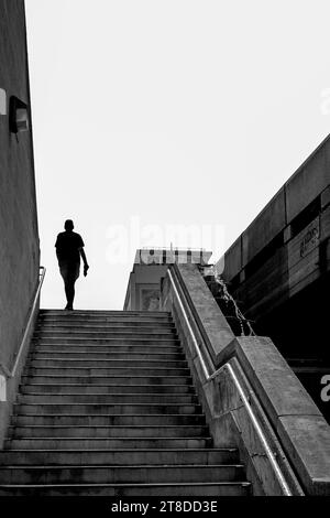 Schwarz-weißes Bild der Silhouette eines Mannes, der draußen eine breite Betontreppe vor hellem Himmel hinaufgeht. Stockfoto