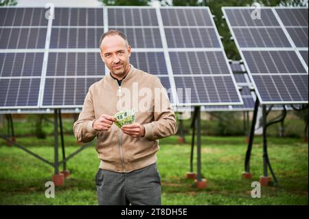 Junge Männer zählen Geld, das er durch Investitionen in alternative Ressourcen erhielt. Glücklicher Mann, der direkt in die Kamera lächelt. Mann, der Euro auf dem Hintergrund des Sonnenkollektors hält. Stockfoto