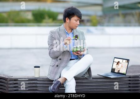 Asiatischer Geschäftsmann genießt Salat, während er Webinar auf Laptop im Freien ansieht Stockfoto