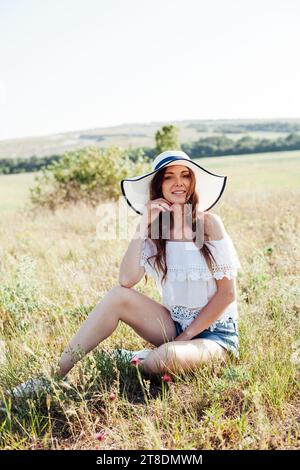 Eine schöne Frau mit Hut sitzt auf trockenem Gras auf einem Feld Stockfoto