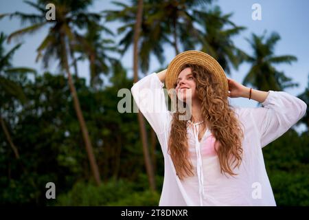 Die fröhliche junge Frau genießt einen tropischen Zufluchtsort zwischen üppigen Palmen, hält sich an ihrem Strohhut fest und genießt die Gelassenheit der Natur mit einer warmen, zufriedenen Atmosphäre Stockfoto