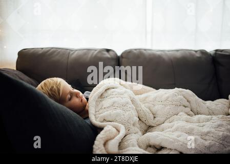 Seitenansicht eines kleinen Jungen, der auf grauer Couch mit weißer Decke schläft Stockfoto