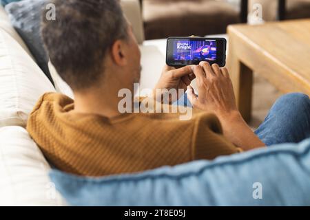 Glücklicher, älterer birassischer Mann, der auf der Couch sitzt und zu Hause Filme auf dem Smartphone ansieht Stockfoto