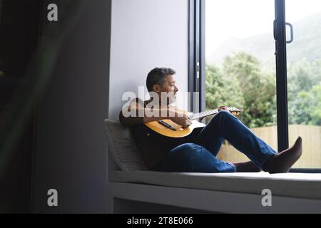 Ein glücklicher Senior-Biracial-Mann, der zu Hause am Fenster Gitarre spielt Stockfoto