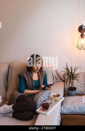 Vertikal, Frau, mitteljung, kaukasisch, kurzes Haar, attraktiv, mit lässiger Kleidung, die ein eBook liest, mit einem Aufguss und ein paar Nüssen. Sie sitzt Stockfoto