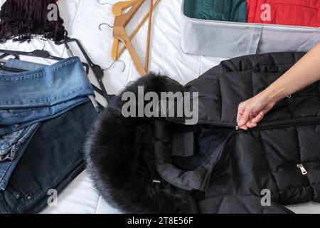 Horizontales Foto, die Hand der Frau schließt den Reißverschluss eines schwarzen Mantels, auf einem Bett, um Winterkleidung in Stoffkisten zu sammeln, zum Jahreswechsel. Stockfoto