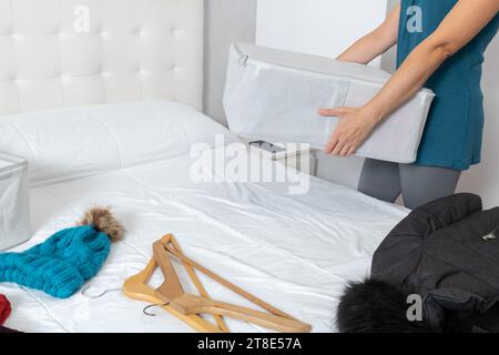 Horizontales Foto, Frau mit einer Kiste Winterkleidung in ihrem Zimmer, für den Jahreswechsel. Es gibt Kleiderbügel und Winterkleidung auf dem Bett. Stockfoto