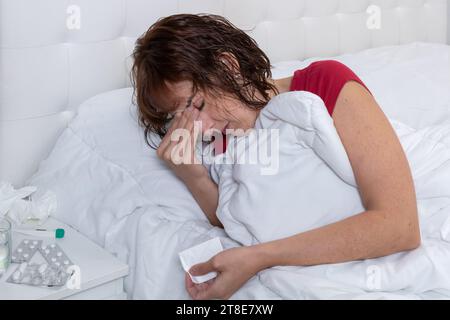 Horizontales Foto kaukasische mittlere Erwachsene Frau, krank, mit Kopfschmerzen, im Bett mit rotem Pyjama und einem Schal in der Hand. Stockfoto