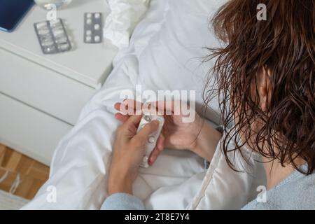Horizontales Foto einer kranken kaukasischen mittelerwachsenen Frau mit langem blauen Pyjama. Nehmen Sie eine Pille aus der Blisterpackung, um das Medikament im Bett einzunehmen. Stockfoto
