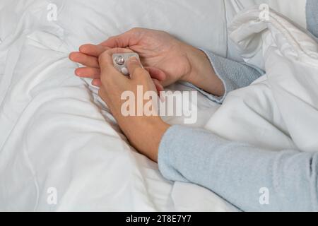 Horizontales Foto einer kranken kaukasischen mittelerwachsenen Frau mit langem blauen Pyjama. Nehmen Sie eine Pille aus der Blisterpackung, um das Medikament im Bett einzunehmen. Stockfoto