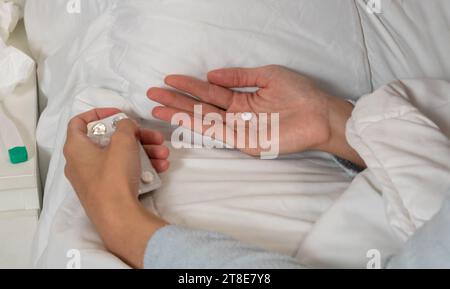 Horizontales Foto einer kranken kaukasischen mittelerwachsenen Frau mit langem blauen Pyjama. Nehmen Sie eine Pille aus der Blisterpackung, um das Medikament im Bett einzunehmen. Stockfoto