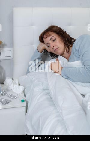 Vertikales Foto, kaukasische mittelerwachsene Frau, mit langem blauen Pyjama, krank im Bett, neben einem Nachttisch mit Medikamenten, einem Thermometer. Stockfoto