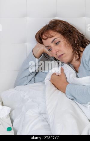 Vertikales Foto, mittelErwachsene kaukasische Frau, mit langem blauen Pyjama, krank im Bett, bedeckt mit einer weißen Decke. Stockfoto