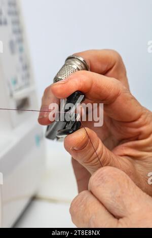 Vertikales Foto, kaukasische Hände einer erwachsenen reifen Schneiderin, mit Fingerhut am Mittelfinger, Schneiden des Garns mit einer Schere auf der Nähmaschine. Stockfoto