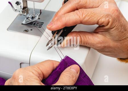 Horizontales Nahaufnahme-Foto, kaukasische Hände einer erwachsenen reifen Näherin, mit Fingerhut am Mittelfinger, Schneiden des Fadens mit einer Schere. Stockfoto