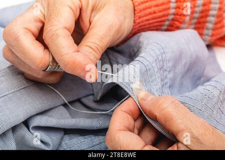 Foto Nahaufnahme extrem horizontal, Hände einer Reifen, erwachsenen kaukasischen Frau, Hand nähen einen Knopf mit Nadel und Fingerhut, auf einem blauen Hemd. Stockfoto