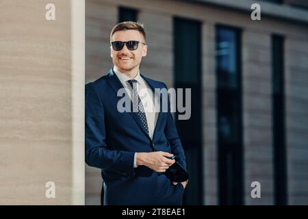 Ein Geschäftsmann in einer Sonnenbrille, der ein Smartphone hält, mit modernen Bürogebäuden im Hintergrund Stockfoto