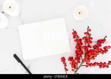 Minimale Weihnachtsdekoration Kerzen und Zweig mit roten Beeren und offenes leeres Notizbuch auf weißem Marmortischhintergrund. Weihnachten, Neujahr, Winte Stockfoto