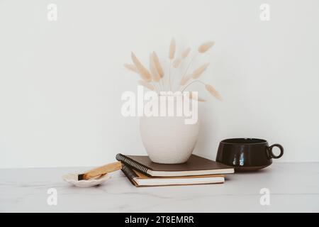 Stilleben von trockenen Lagurus-Blüten in Vase, Tasse Tee, Notizbücher und palo santo auf weißem Tisch. Kopierbereich. Vorderansicht Stockfoto