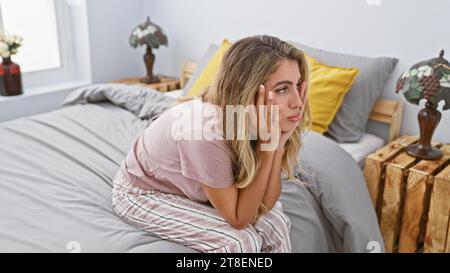 Erschöpfte junge blonde Frau, gestresst über die Probleme des Lebens, sitzt auf ihrem Bett in einem düsteren Schlafzimmer und unterstreicht den Schmerz der Angst Stockfoto