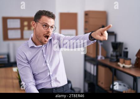 Junger hispanischer Mann im Büro, der mit dem Finger überrascht vorne zeigt, offener Mund, verwunderter Ausdruck, etwas auf der Vorderseite Stockfoto