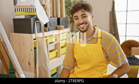Selbstbewusster, lächelnder junger arabischer Mann, Künstler aus Leidenschaft, sitzt auf einem Kunststudio-Stuhl, taucht in Gemälde ein Stockfoto