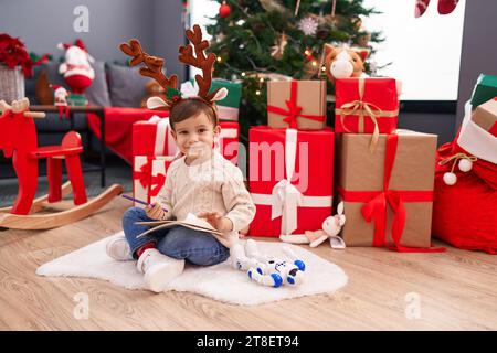 Ein entzückender hispanischer Junge, der den weihnachtsmann-Brief schreibt, sitzt zu Hause am weihnachtsbaum auf dem Boden Stockfoto