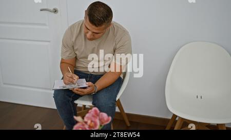 Ernsthafter lateinmann, entspannt und konzentriert, sitzt im Stuhl und macht Notizen. Innenporträt eines jungen hispanischen Mannes, der eine Checkliste für das Bewerbungsgespräch auf Clip schreibt Stockfoto