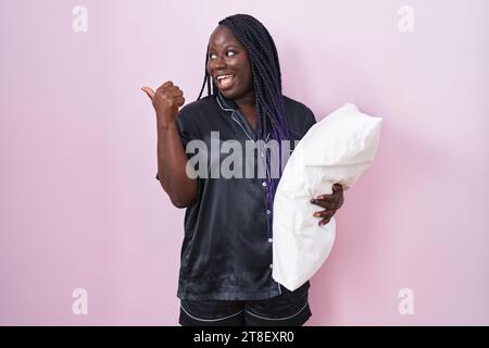 Junge afrikanische Frau, die einen Pijama trägt, das Kissen umarmt, lächelnd mit glücklichem Gesicht, das aussieht und mit dem Daumen nach oben zur Seite zeigt. Stockfoto