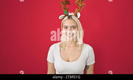 Junge schöne hispanische Frau lächelt selbstbewusst mit Rentier-weihnachtshut über isoliertem roten Hintergrund Stockfoto