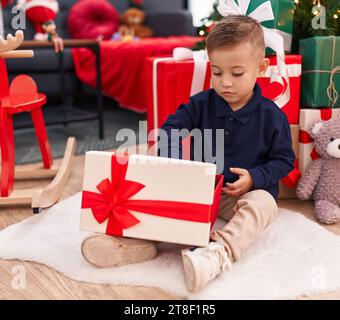 Entzückender hispanischer Junge, der zu Hause am weihnachtsbaum sitzt und ein Geschenk auspackt Stockfoto